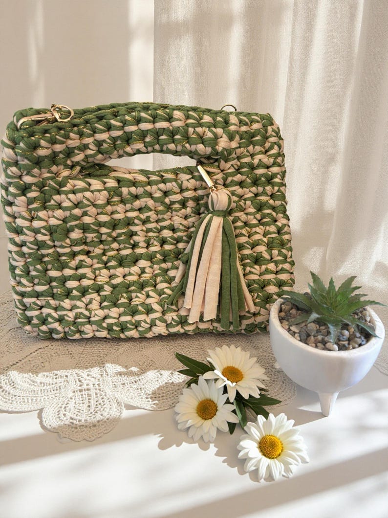 K&ouml;nnte beinhalten: Handgefertigte gr&uuml;ne und cremefarbene H&auml;keltasche mit rechteckiger Form und Griff. Die Tasche hat einen goldfarbenen Kettenriemen und einen Quastenanh&auml;nger. G&auml;nsebl&uuml;mchen und eine kleine Sukkulente in einem wei&szlig;en Topf sind ebenfalls im Bild.