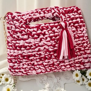 K&ouml;nnte beinhalten: Handgefertigte, rechteckige Handtasche in einem rot-rosa Webmuster. Die Tasche hat einen Griff, einen goldenen Kettenriemen und einen Quastenanh&auml;nger. Die Tasche ist etwa 30 cm breit und 20 cm hoch.