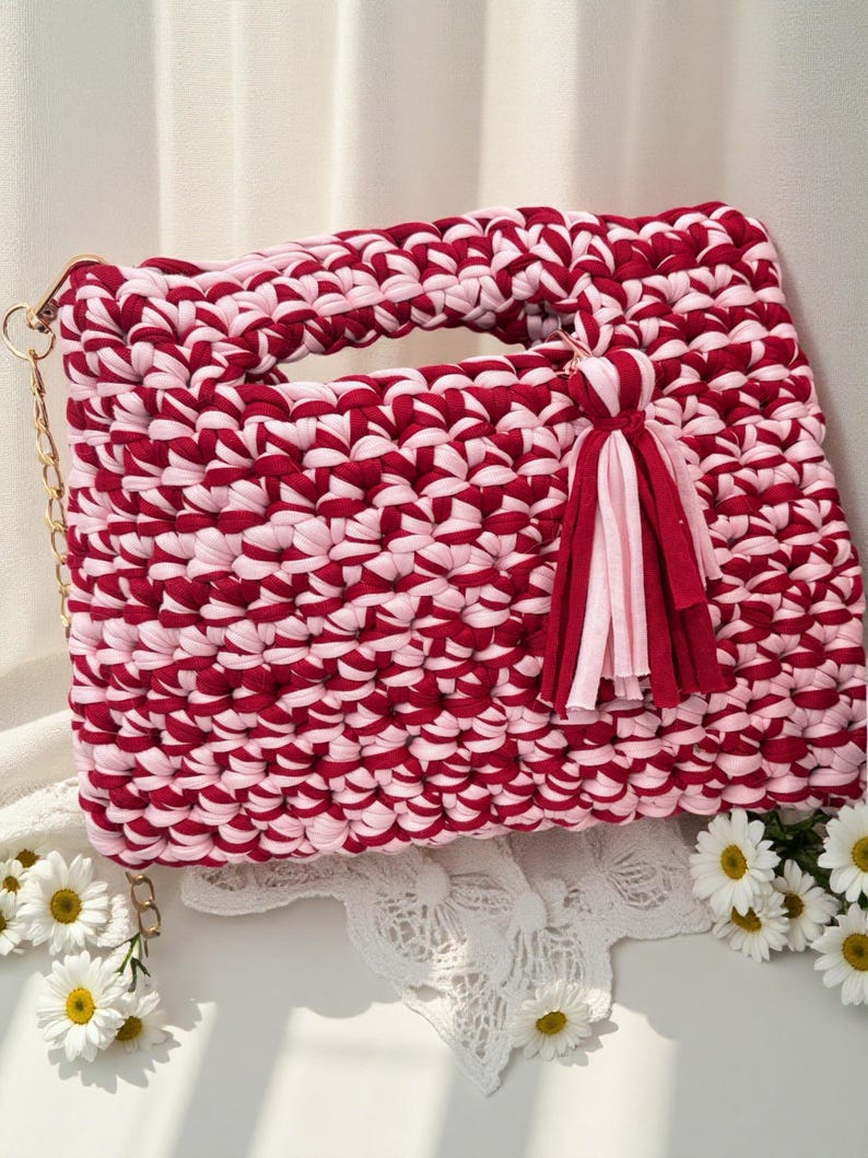 K&ouml;nnte beinhalten: Handgefertigte, rechteckige Handtasche in einem rot-rosa Webmuster. Die Tasche hat einen Griff, einen goldenen Kettenriemen und einen Quastenanh&auml;nger. Die Tasche ist etwa 30 cm breit und 20 cm hoch.