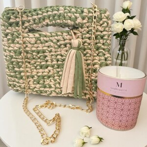 K&ouml;nnte beinhalten: Handgefertigte gr&uuml;ne und cremefarbene H&auml;keltasche mit goldener Kette und Quaste. Die Tasche ist quadratisch mit Griff und dekorativer Quaste. Eine rosa Kerze mit goldenen Akzenten und wei&szlig;e Rosen in einer Vase sind ebenfalls vorhanden.