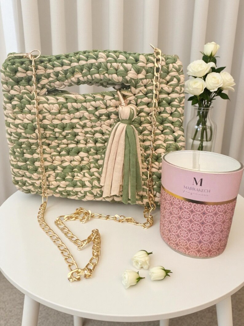 K&ouml;nnte beinhalten: Handgefertigte gr&uuml;ne und cremefarbene H&auml;keltasche mit goldener Kette und Quaste. Die Tasche ist quadratisch mit Griff und dekorativer Quaste. Eine rosa Kerze mit goldenen Akzenten und wei&szlig;e Rosen in einer Vase sind ebenfalls vorhanden.