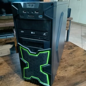 Pode incluir: Uma torre de computador preta com um painel frontal que apresenta um design em forma de X com contornos em verde brilhante. A parte frontal inclui uma unidade de disco ótico LG, várias portas USB, entradas de áudio e um botão de energia.