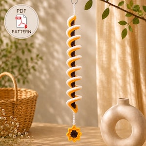 Könnte beinhalten: Ein gehäkeltes Spiral-Hängedekor mit dunkelbrauner Mitte sowie gelben und weißen Rändern, aufgehängt an einer weißen Schnur. Eine kleine gelbe Sonnenblume mit dunkelbraunem Kern hängt unten. Enthält ein PDF PATTERN Etikett.