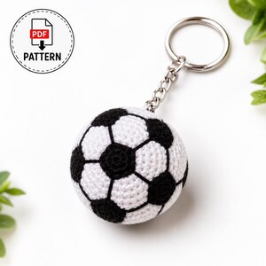 Op de afbeelding: Een gehaakte voetbal-sleutelhanger met een klassiek zwart-wit zeshoekig patroon, bevestigd aan een zilveren metalen ring met een korte ketting. Een cirkelvormig embleem linksboven toont de tekst PDF PATTERN met een downloadpictogram.