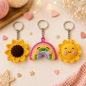 Könnte beinhalten: Drei gehäkelte Schlüsselanhänger: eine gelbe Sonnenblume, ein Regenbogen und eine lächelnde Sonne. Jeder Schlüsselanhänger hat eine silberne Kette und einen Ring. Die Sonnenblume hat eine braune Mitte. Der Regenbogen hat rosa, grüne, blaue und gelbe Bögen. Die Sonne ist gelb mit einem lächelnden Gesicht.
