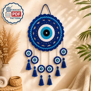 Puede incluir: Un tapiz circular de ganchillo con un motivo central de ojo turco en azul marino, azul claro y blanco con un centro negro. Seis adornos de ojo más pequeños cuelgan de hilos de cuentas blancas, cada uno terminado con una borla azul marino.