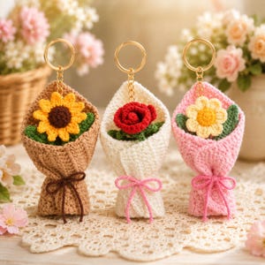 May include: Three crochet flower keychains featuring a sunflower in a tan wrap, a red rose in a white wrap, and a yellow daisy in a pink wrap. Each bouquet is tied with a small matching yarn bow and attached to a gold-toned metal keyring.