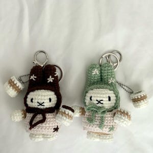 May include: Two crochet rabbit keychains featuring white faces with black X-shaped mouths and eyes. One rabbit wears a dark brown hood with white flower accents, while the other wears a sage green hood with white flower accents. Both hold small cups.