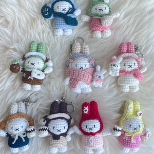 May include: A collection of handmade crochet bunny keychains in various colors. Each bunny has a unique outfit, including hats, scarves, and dresses. The keychains are white, blue, green, pink, brown, and yellow, with black embroidered facial features.