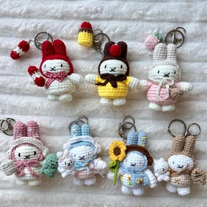 Peut inclure: Une collection de porte-clés lapins crochetés à la main, dans différentes couleurs et tenues. Chaque lapin a un design unique, avec des chapeaux, des écharpes et des accessoires comme un tournesol ou un petit gâteau. Les porte-clés sont fixés à des anneaux métalliques argentés.
