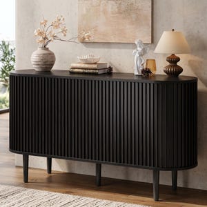 May include: A black sideboard with a rounded, fluted exterior and four tapered legs. The top surface holds a ribbed vase with dried flowers, a white bowl on two books, a white angel figurine, a small amber glass, and a table lamp with a beige shade.