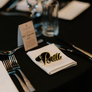 May include: A formal table setting with a black tablecloth, cutlery, and glassware. A folded white napkin holds a name card reading "Pritt" in gold and black. A menu card is also visible.