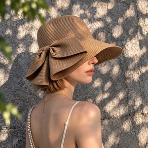 May include: A tan straw sun hat featuring a wide, downward-sloping brim and a large, pleated bow detail on the side. The hat has a ribbed texture throughout the material and is worn with a white camisole top featuring thin, pearl-adorned straps.