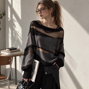 May include: A long-sleeved jumper featuring horizontal stripes in black and charcoal grey. The knit design incorporates alternating opaque sections and sheer, semi-transparent panels. The garment has a wide, relaxed neckline and ribbed cuffs.