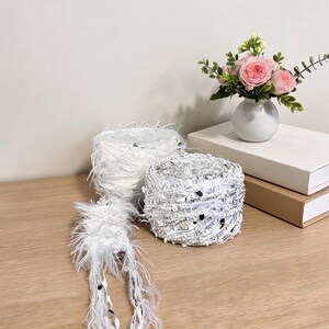 May include: Two skeins of white novelty yarn with a fuzzy, eyelash-like texture and embedded reflective silver sequins. The yarn is wound into compact, cylindrical balls, with a loose strand extending forward, showcasing the soft, feathery fibers.