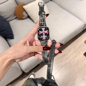 May include: A smartwatch featuring a dark gray textured leather strap adorned with silver-toned cross-shaped metal embellishments. The watch face displays a digital clock reading 17:01 with a prominent cross graphic and intricate text details.