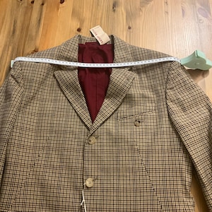 Puede incluir: Una blazer a cuadros marr&oacute;n y beige con forro burdeos. La blazer tiene un cierre cl&aacute;sico de dos botones y solapas con muescas. Una cinta m&eacute;trica est&aacute; colocada sobre los hombros. La etiqueta dice "antiquit&eacute;s".