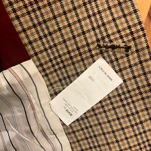 Puede incluir: Primer plano de una blazer a cuadros beige y negro. La blazer tiene un forro burdeos y una etiqueta blanca con el texto "MADE IN ITALY". El forro interior es blanco con rayas rojas y negras.