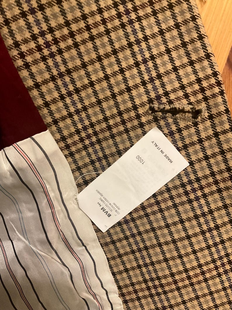 Puede incluir: Primer plano de una blazer a cuadros beige y negro. La blazer tiene un forro burdeos y una etiqueta blanca con el texto "MADE IN ITALY". El forro interior es blanco con rayas rojas y negras.