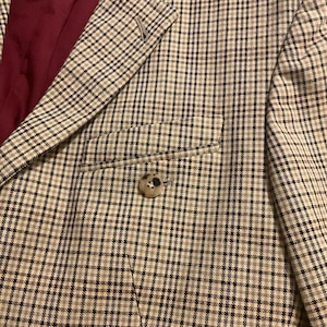 Puede incluir: Primer plano de una blazer a cuadros beige y negro con cierre de un solo bot&oacute;n. La blazer tiene un forro burdeos y un bolsillo en la parte delantera. La tela tiene un sutil patr&oacute;n de cuadr&iacute;cula azul.