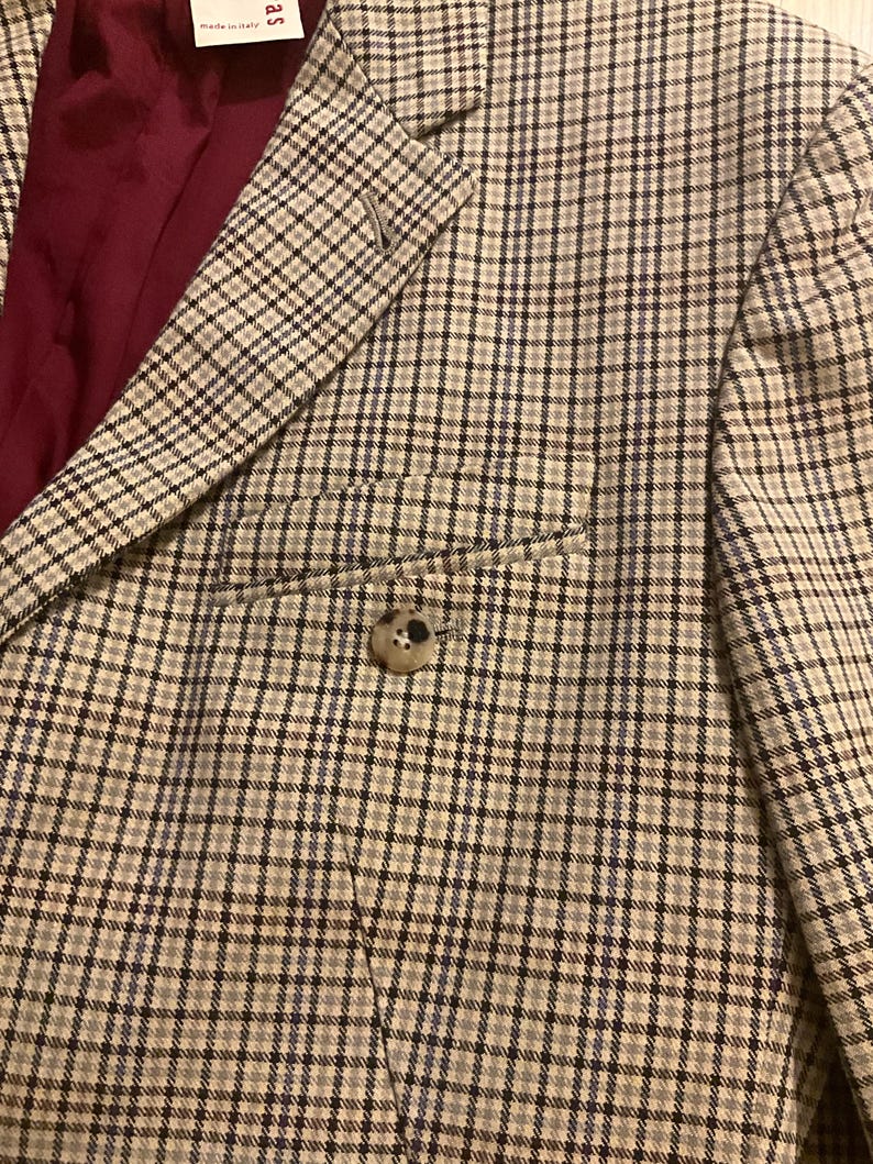 Puede incluir: Primer plano de una blazer a cuadros beige y negro con cierre de un solo bot&oacute;n. La blazer tiene un forro burdeos y un bolsillo en la parte delantera. La tela tiene un sutil patr&oacute;n de cuadr&iacute;cula azul.
