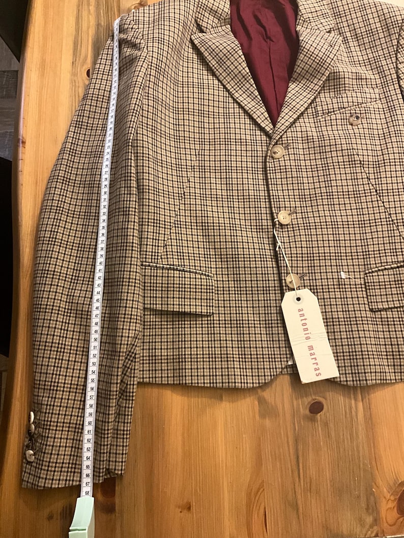 Puede incluir: Una blazer a cuadros beige y negro con forro burdeos. La blazer tiene dos botones y una etiqueta que dice "antonio marras". Una cinta m&eacute;trica est&aacute; al lado de la blazer, mostrando una longitud de aproximadamente 173 cm.