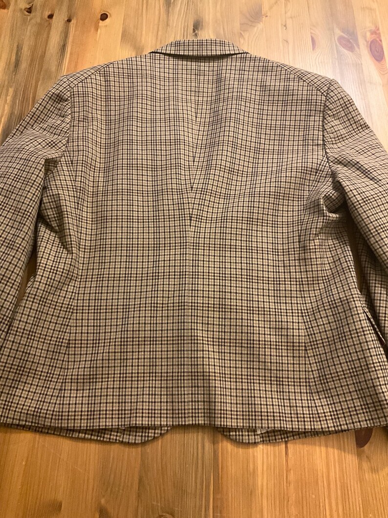 Puede incluir: Un blazer a cuadros marr&oacute;n y negro con solapas y cierre de dos botones. Se ve la parte trasera del blazer, mostrando un corte entallado y una abertura central. La tela tiene un patr&oacute;n tejido cl&aacute;sico.