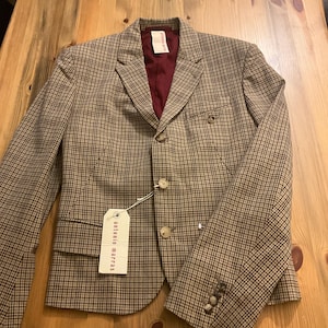 Puede incluir: Una blazer a cuadros marr&oacute;n y negro con forro burdeos. La blazer tiene dos botones, solapa con muescas y una etiqueta que dice "antonio marras". La blazer est&aacute; sobre una superficie de madera.