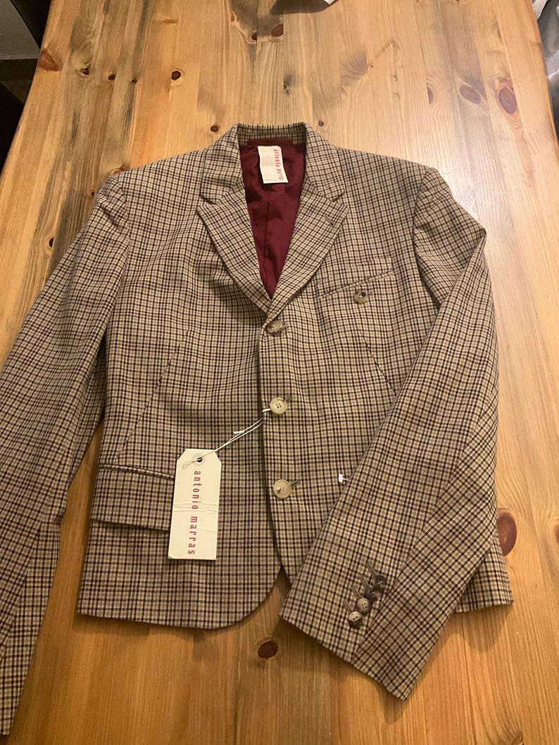 Puede incluir: Una blazer a cuadros marr&oacute;n y negro con forro burdeos. La blazer tiene dos botones, solapa con muescas y una etiqueta que dice "antonio marras". La blazer est&aacute; sobre una superficie de madera.