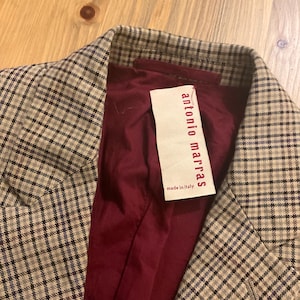 Puede incluir: Primer plano de una blazer con estampado de cuadros en tonos beige, gris y azul. El forro interior es de color burdeos oscuro. Una etiqueta dice "antonio marras made in italy".