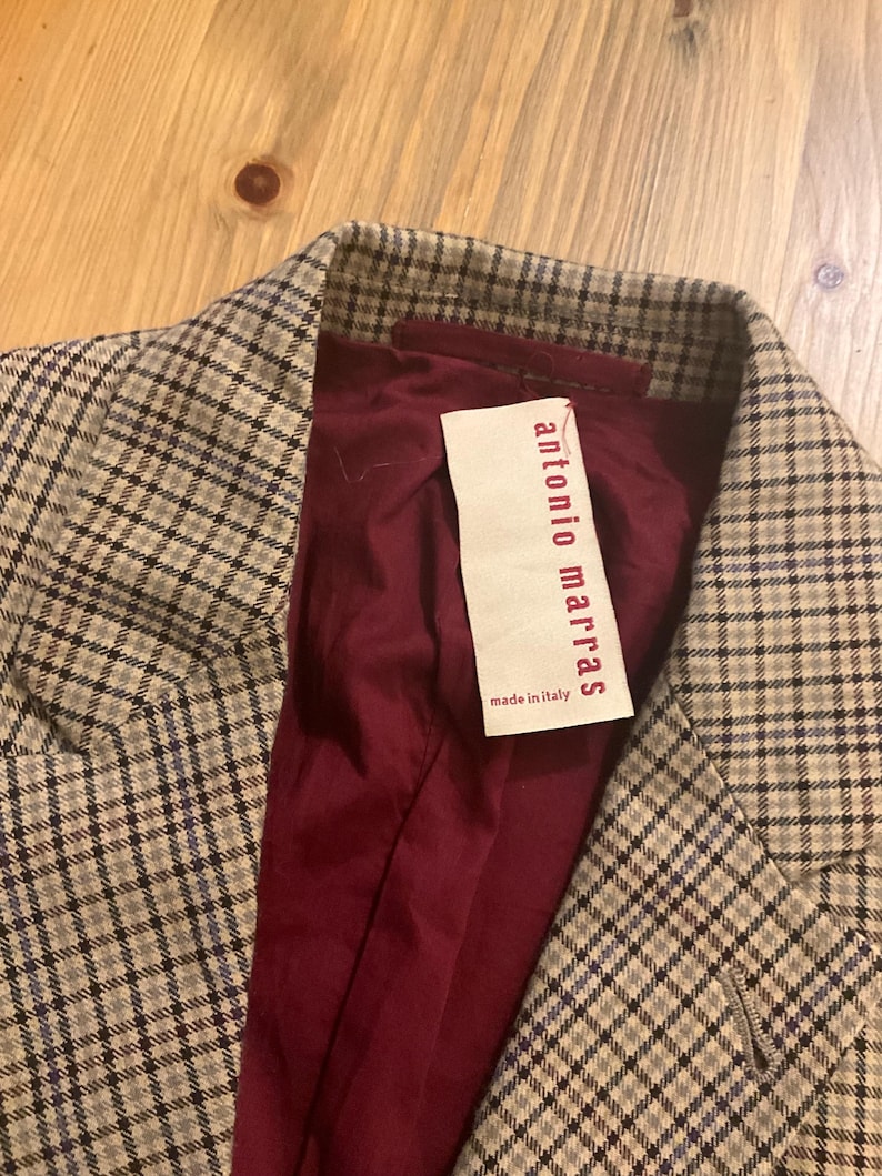 Puede incluir: Primer plano de una blazer con estampado de cuadros en tonos beige, gris y azul. El forro interior es de color burdeos oscuro. Una etiqueta dice "antonio marras made in italy".