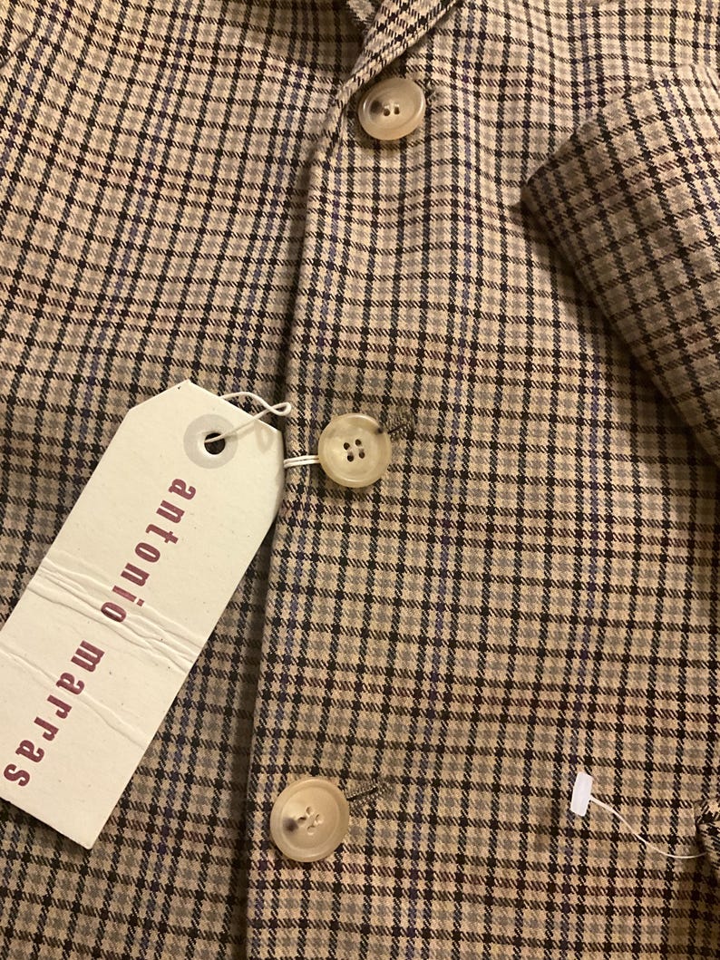 Puede incluir: Primer plano de una blazer a cuadros marr&oacute;n y beige con tres botones color crema. La blazer tiene una etiqueta que dice "antonio marras". La tela tiene un sutil patr&oacute;n de cuadros azules.