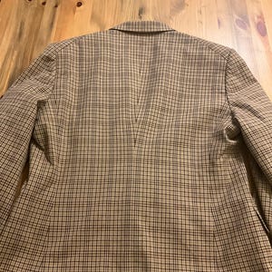 Puede incluir: Una blazer a cuadros marr&oacute;n y beige, vista desde atr&aacute;s, est&aacute; sobre una superficie de madera. La blazer tiene un cuello con solapas y un dise&ntilde;o cl&aacute;sico. El patr&oacute;n de cuadros es un peque&ntilde;o cuadriculado repetido.