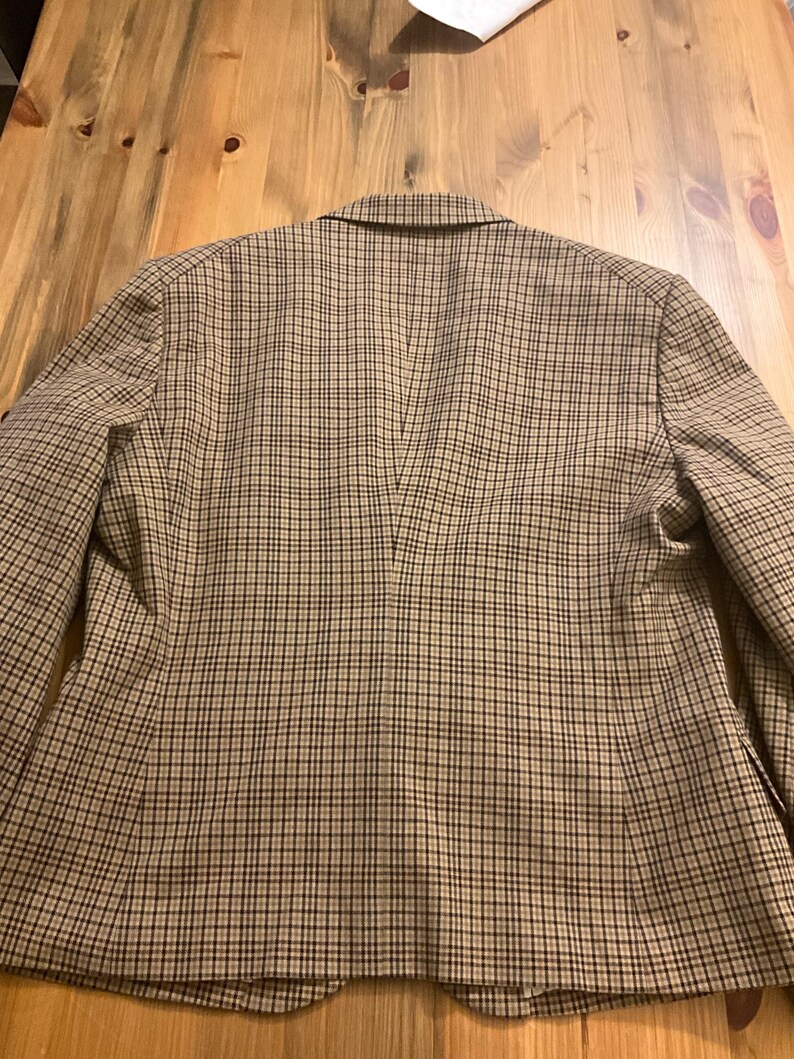 Puede incluir: Una blazer a cuadros marr&oacute;n y beige, vista desde atr&aacute;s, est&aacute; sobre una superficie de madera. La blazer tiene un cuello con solapas y un dise&ntilde;o cl&aacute;sico. El patr&oacute;n de cuadros es un peque&ntilde;o cuadriculado repetido.