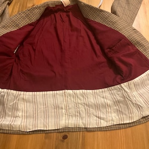 Puede incluir: Un blazer a cuadros marr&oacute;n y beige con forro burdeos y ribete a rayas blancas y marrones. El blazer tiene un dise&ntilde;o cl&aacute;sico y parece sin usar. Una etiqueta es visible cerca del cuello.