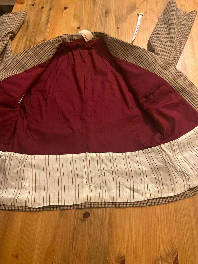 Puede incluir: Un blazer a cuadros marr&oacute;n y beige con forro burdeos y ribete a rayas blancas y marrones. El blazer tiene un dise&ntilde;o cl&aacute;sico y parece sin usar. Una etiqueta es visible cerca del cuello.