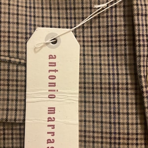 Puede incluir: Primer plano de una tela a cuadros marr&oacute;n y gris con una etiqueta blanca adjunta. La etiqueta tiene las palabras "antonio marras" impresas en color rosa oscuro. La tela tiene un patr&oacute;n de cuadros cl&aacute;sico.
