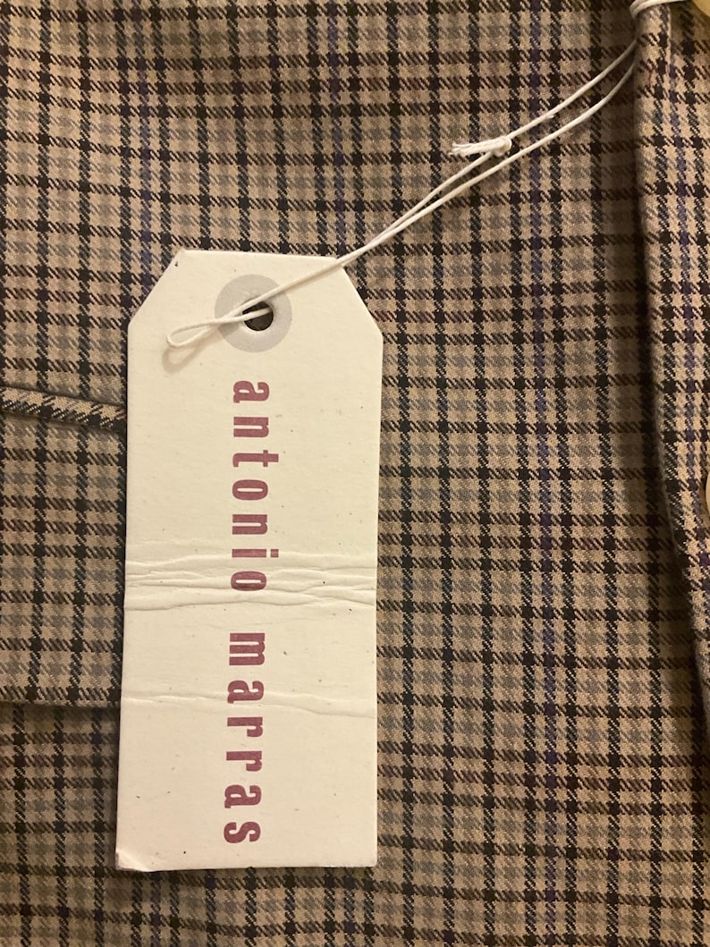 Puede incluir: Primer plano de una tela a cuadros marr&oacute;n y gris con una etiqueta blanca adjunta. La etiqueta tiene las palabras "antonio marras" impresas en color rosa oscuro. La tela tiene un patr&oacute;n de cuadros cl&aacute;sico.