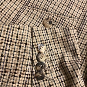 Puede incluir: Primer plano de una blazer a cuadros beige y azul. La blazer tiene cuatro botones en la manga y uno en la solapa. Los botones son de color beige y azul veteado. La tela tiene un patr&oacute;n sutil.