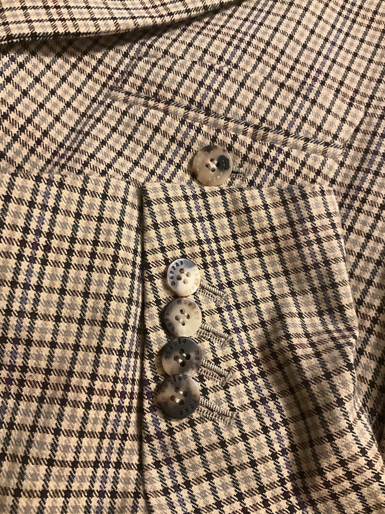 Puede incluir: Primer plano de una blazer a cuadros beige y azul. La blazer tiene cuatro botones en la manga y uno en la solapa. Los botones son de color beige y azul veteado. La tela tiene un patr&oacute;n sutil.
