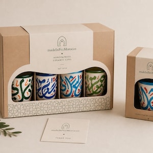 May include: A set of four handpainted ceramic cups in a brown cardboard box labeled madeInFezMorocco, featuring colorful Arabic calligraphy designs in blue, green, and red. A separate smaller box and a thank you card are also visible in the display.
