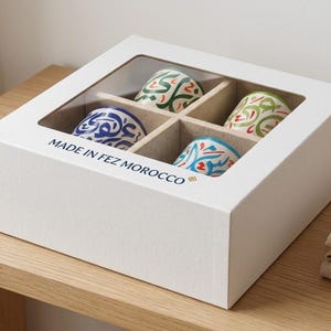 May include: A white square cardboard box with a clear window containing four ceramic cups. Each cup features colourful Arabic calligraphy patterns in blue, green, and red. The box is printed with the text MADE IN FEZ MOROCCO on the front side.