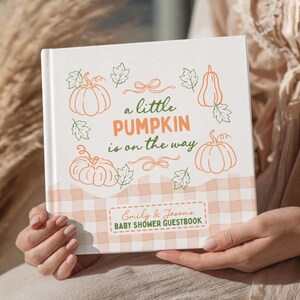 May include: A square hardback guestbook with a white background featuring orange pumpkin illustrations, green leaf outlines, and the text 'a little PUMPKIN is on the way'. The bottom has a beige gingham pattern and a label reading 'Emily & Jason's BABY SHOWER GUESTBOOK'.