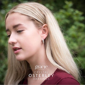 Può includere: Una donna con lunghi capelli biondi indossa orecchini di cristallo. Gli orecchini sono piccoli cerchi con più cristalli. L'immagine include il testo "gracie LONDON CRYSTAL OSTERLEY EARRINGS". La donna indossa un top bordeaux.