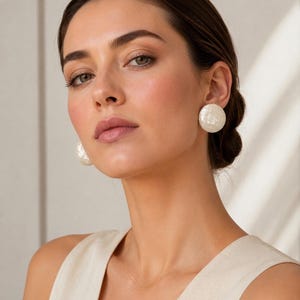 Può includere: Una donna indossa grandi orecchini rotondi di perle bianche. Gli orecchini hanno una superficie strutturata. La donna indossa un top senza maniche. Lo sfondo è neutro.