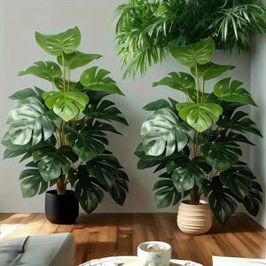 May include: Two artificial monstera plants with large, split green leaves. One plant is in a smooth black pot, and the other is in a ribbed beige pot. Both plants feature tall, upright stems and lush foliage, positioned side by side in a room.