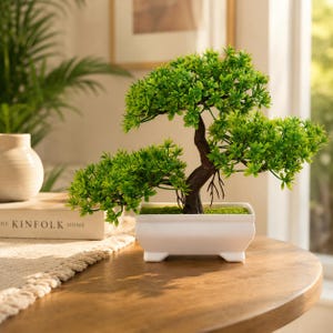 Könnte beinhalten: Ein kleiner künstlicher Bonsai-Baum mit leuchtend grünem Blattwerk und einem dunkelbraunen, strukturierten Stamm. Der Baum ist in einen rechteckigen, weißen Keramiktopf mit vier kleinen Füßen gepflanzt, dessen Erdoberfläche mit grünem Moos bedeckt ist.