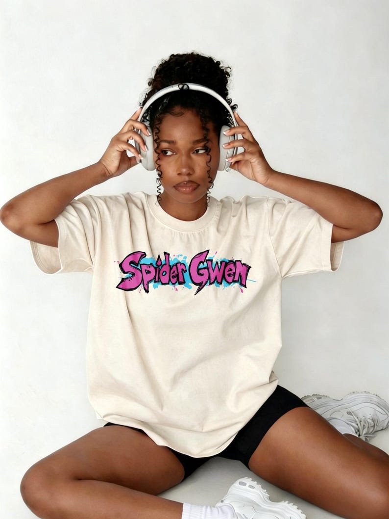May include: A cream-coloured short-sleeve t-shirt featuring the text "Spider Gwen" in a stylised pink and black graffiti-inspired font with blue paint splatter accents across the chest. The shirt has a crew neckline and a relaxed fit.