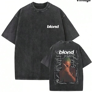May include: A charcoal gray short-sleeve t-shirt with a washed finish. The front features the word "blond" in white. The back displays a graphic of a person covering their face with text including "blond," "Godspeed," and "Facebook Story."