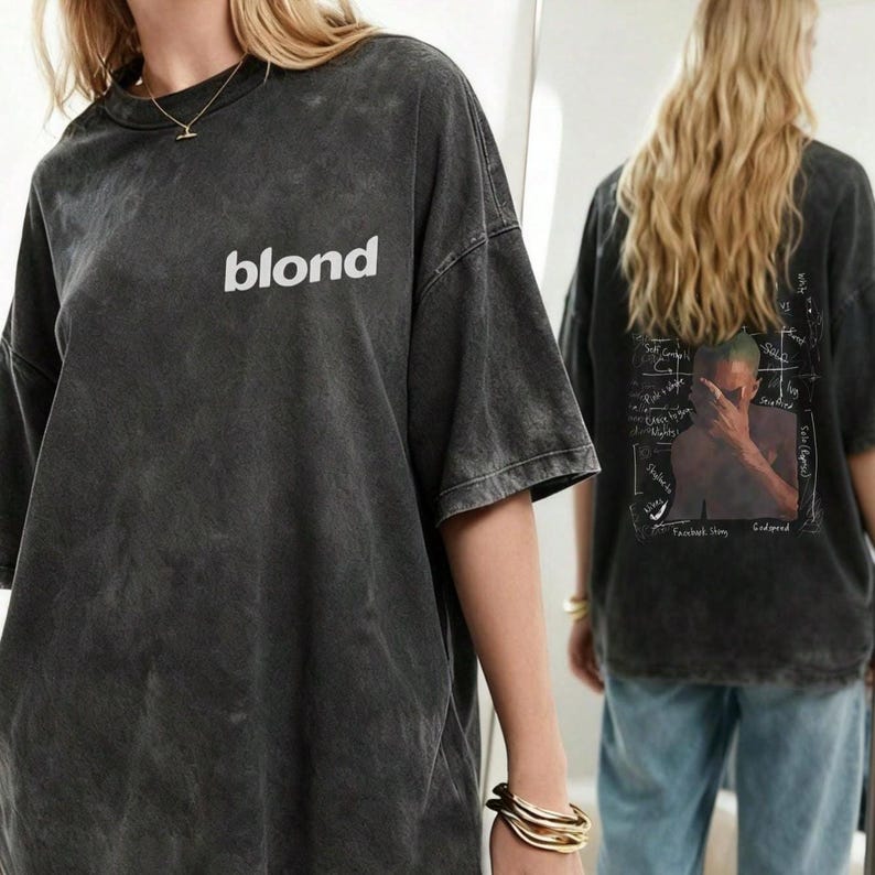 May include: A charcoal grey acid-wash t-shirt featuring the word blond in white text on the front chest. The back of the shirt displays a graphic of a person covering their face with their hand, surrounded by handwritten-style text and markings.